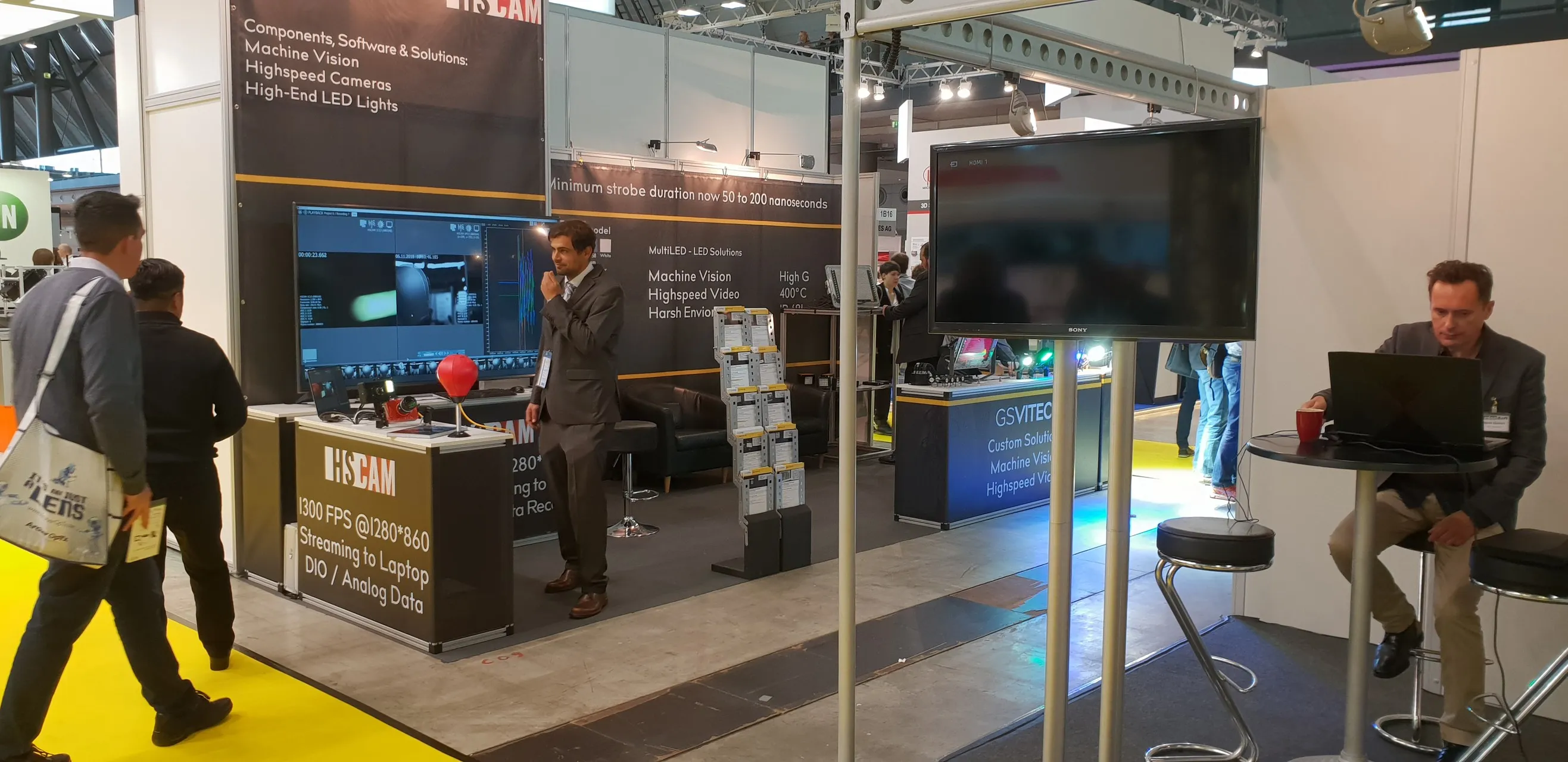 Wide shot of HSCAM's own booth at the Messe Vision 2018 trade fair, showing the punching-ball rig and an HSCAM representative beside it.